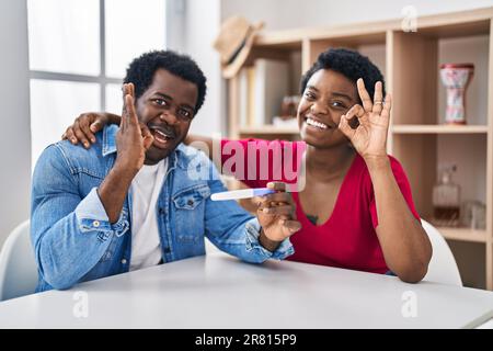 Young african american couple holding pregnancy test result doing ok sign with fingers, smiling friendly gesturing excellent symbol Stock Photo