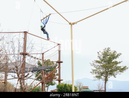 Teenage teen girl bungee flying in rope amusement park. Climbing harness equipment, green sports safety helmet. Hanging obstacle course. Kids children Stock Photo