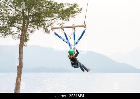 Teenage teen girl bungee flying in rope amusement park. Climbing harness equipment, green sports safety helmet. Hanging obstacle course. Kids children Stock Photo