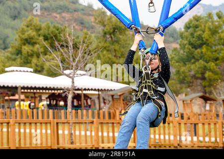 Teenage teen girl bungee flying in rope amusement park. Climbing harness equipment, green sports safety helmet. Hanging obstacle course. Kids children Stock Photo