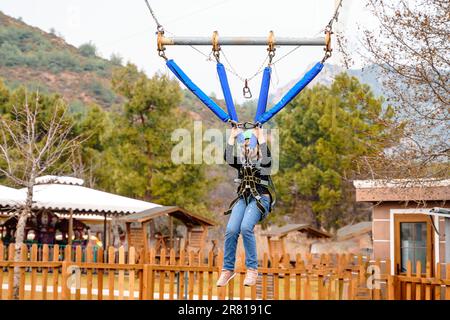 Teenage teen girl bungee flying in rope amusement park. Climbing harness equipment, green sports safety helmet. Hanging obstacle course. Kids children Stock Photo