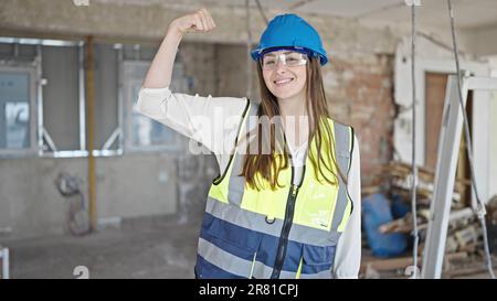 Young beautiful hispanic woman builder smiling confident doing strong gesture with arm at construction site Stock Photo