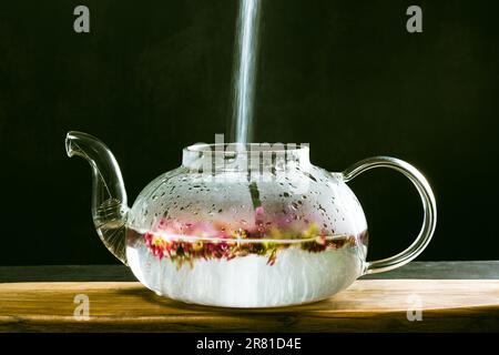 A beautiful glass teapot for brewing tea stands on a wooden board on a dark background, the teapot is transparent. Hot water is poured into kettle, cl Stock Photo