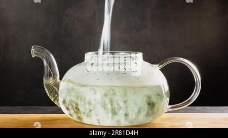 A beautiful glass teapot for brewing tea stands on a wooden board on a dark background, the teapot is transparent. Hot water is poured into the kettle Stock Photo