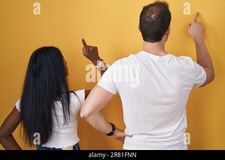 Interracial couple standing over yellow background posing backwards pointing ahead with finger hand Stock Photo