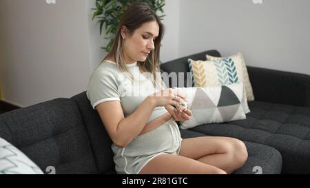 Young pregnant woman smiling confident applying lotion on belly at home Stock Photo