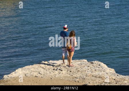 The back of a young couple standing on the edge of the cliff watching the blue sea water below. Stock Photo