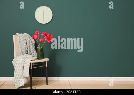 Vase with red carnations and blanket on chair near dark green wall Stock Photo