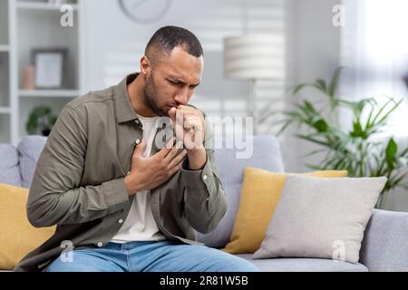 Young African American man sitting on the couch at home and coughing. Suffers from an attack of asthma, allergies. He holds his chest, covers his mouth with his hand. Stock Photo