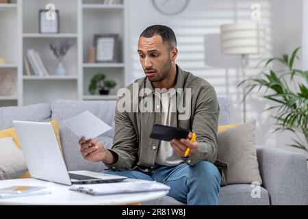 Young african american freelancer businessman working remotely from home with laptop and documents. Worriedly holding a calculator and papers, has financial problems. Stock Photo