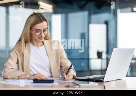 Young female accountant, business woman working in office at laptop and with documents, counting on calculator, paperwork. Stock Photo