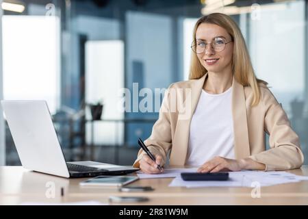 Portrait of a young female student, intern. Sitting in the office at the table, studying online from a laptop, looking at the camera with a smile. Stock Photo