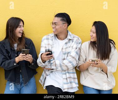 Three smiling young friends browsing social media with smart phone on yellow wall background Stock Photo