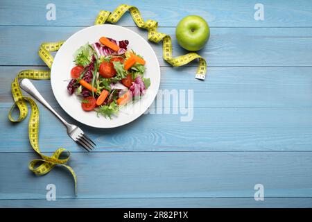 Plate of fresh vegetable salad, apple and measuring tape on light blue wooden table, flat lay with space for text. Healthy diet concept Stock Photo