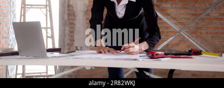Young caucasian architect woman looking laptop and working drawing blueprint for planning building house on desk, engineer thinking and sketching blue Stock Photo