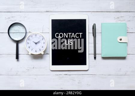 Top view of magnifying glass,table clock,pen,notebook and tablet computer written with ' CONTACT US' on white wooden background. Stock Photo
