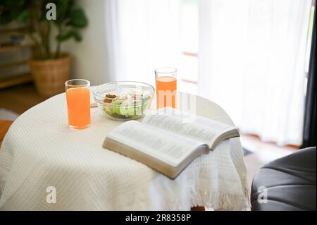 Close-up image of an opened book, a salad bowl, and two glasses of orange juice are on a table in a minimalist dining room. Stock Photo