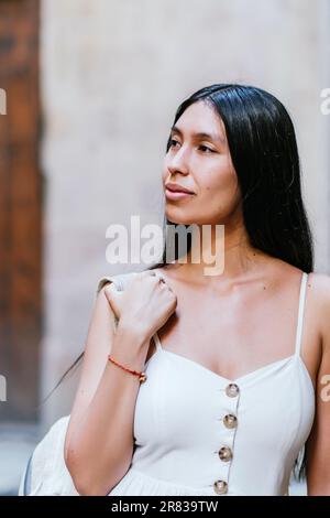 Young Latin American woman looking away and thinking while adjusting backpack on shoulder on blurred background of street of Barcelona, Spain Stock Photo