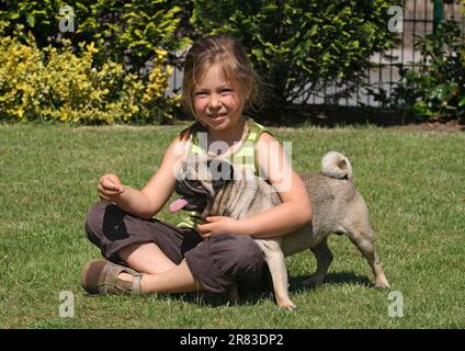 Little girl sits with male pug, 1 year old in the garden on the lawn and gives the dog a treat Stock Photo