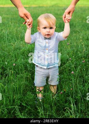 Baby child on the green grass in summer park. Parents hand and child. Perfect family holding hands, adopted child being supported by loving parents. Stock Photo