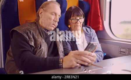 Elderly couple travel by train, the senior is holding a mobile phone in her hand, both are looking at the smartphone and talking with each other Stock Photo