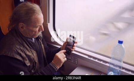 Elderly man traveling by train and taking pictures of the landscape through the window using a smartphone Stock Photo