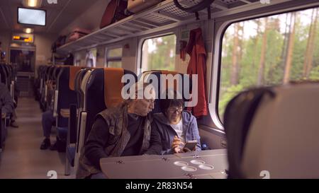 Elderly couple travel by train, the lady is holding a mobile phone in her hand, both are looking at the smartphone and talking with each other Stock Photo