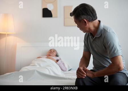 Senior man sitting on bed of sick wife sleeping in bed Stock Photo