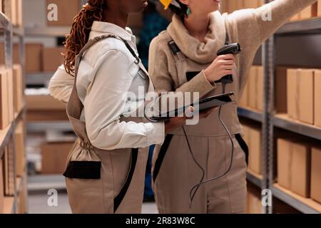 Storehouse colleagues reaching for cardboard box on shelf to scan barcode. Warehouse operators managing received parcels registration with bar code scanner and digital tablet software Stock Photo