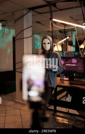 Cybercriminal in anonymous mask recording ransomware video, making online threats on live broadcast. Dangerous spy breaking into server with virus encryption, committing internet fraud. Stock Photo