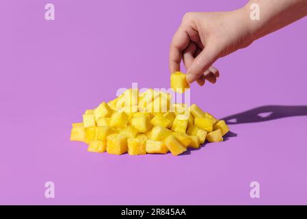 Woman hand taking a piece of pineapple from a pile minimalist on a purple background. Fresh pineapple sliced in small pieces, in bright light on a vib Stock Photo