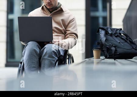 Young man with disability sitting in wheelchair and using laptop outdoors Stock Photo