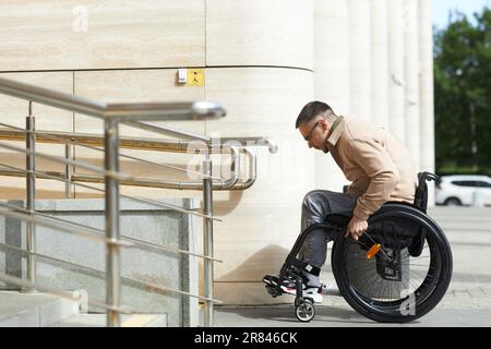 Side view of young man sitting in wheelchair using ramp to move up of building Stock Photo