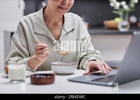 Closeup of smiling senior woman using laptop and working while enjoying breakfast at home Stock Photo