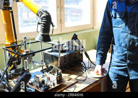 A male working electrician in a robe professionally works on a table for repair in a workshop for remoting electronics and manufacturing parts and spa Stock Photo