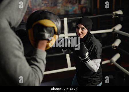 Young Muslim woman in hijab, activewear and leather boxing gloves looking at her trainer with defense pads on hands during training Stock Photo
