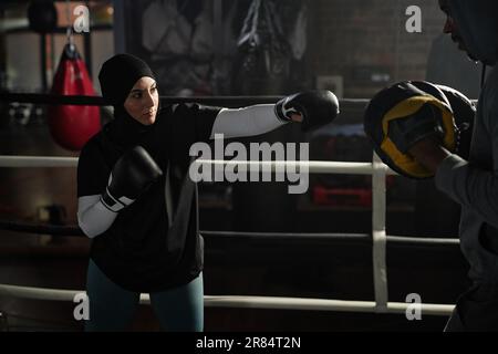 Young Muslim female boxer hitting boxing pads on hands of her trainer while standing in front of him during training on rink in sports club Stock Photo