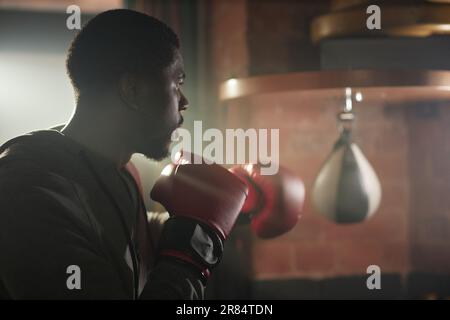 Side view of young African American sportsman in boxing gloves standing in front of camera ready to hit sandbag during training in gym Stock Photo