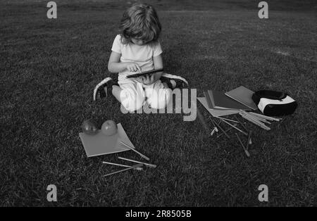 Kids study exam outside. Funny little student boy with tablet, Sit on lawn in park, studying at School backyard. Stock Photo