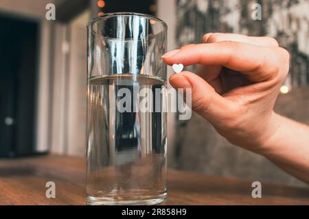 White pill in hand and glass of water. Heart shape pill in hand. Medical treatment. Painkiller from unhappy love. Therapy prescription. Healthcare. Stock Photo