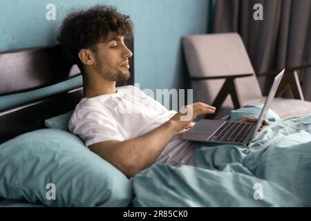 Job in bedroom, social networks, surfing, browsing on internet. Serious young man woke up, sits on bed, works on laptop in interior Stock Photo