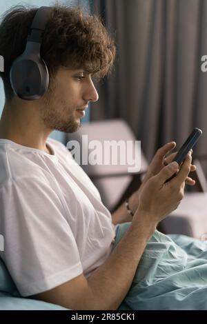Technologies for leisure. Young arab man relaxing in bedroom in wireless headphones using smartphone in bed, listening music Stock Photo