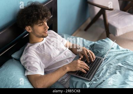 Job in bedroom, social networks, surfing, browsing on internet. Serious young man woke up, sits on bed, works on laptop in interior. Stock Photo