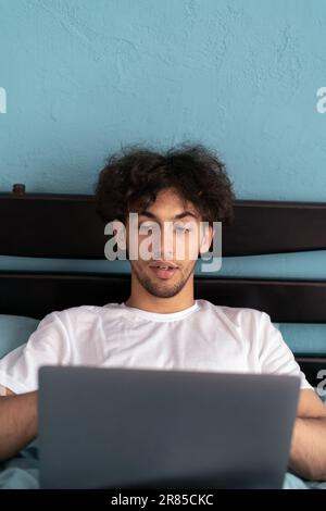 Portrait of arabic guy in weekend morning, working on laptop writing check, wearing nightwear sleep wear t-shirt checkered pants and sitting on bed in Stock Photo