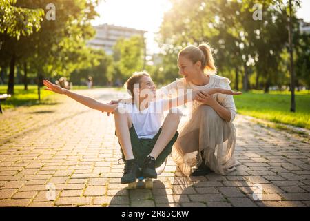 Mother having fun with her son in park on sunny day. Boy is ridding skateboard and his mother is pushing him. Stock Photo