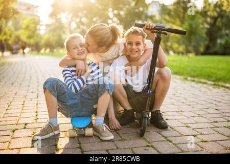 Joyful mother having fun with her two sons in park on sunny day. They are ridding skateboard and scooter. Stock Photo