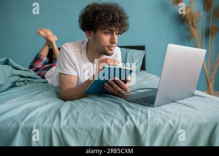 Young arab guy student studying virtual course, learning distance class taking web training, looking at laptop computer, writing notes sitting on bed Stock Photo