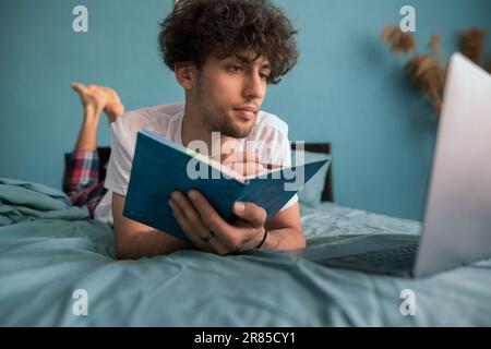 Curly young eastern guy studying in bed, using laptop, watching course on Internet or webinar, taking notes, copy space Stock Photo