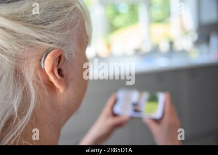 Close Up Of Senior Woman Wearing Behind The Ear Hearing Device Or Aid Sreaming From Mobile Phone At Home Stock Photo