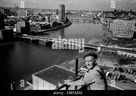 London Skyline 1970s. Irish Navvie Navvy an Irish labourer, a construction worker holding hammer at the top of and working on a Nine Elms tower block. London cityscape view, the  River Thames, Vauxhall Cross below and Waterloo in the distance. London, England 1974. HOMER SYKES Stock Photo